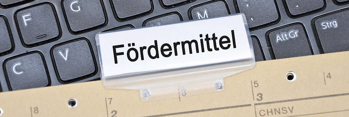 Symbolbild Fördermittel: Registerkarte mit Aufschrift ‚Fördermittel‘ auf Tastatur und Aktenregister – digitale und analoge Antragsunterlagen