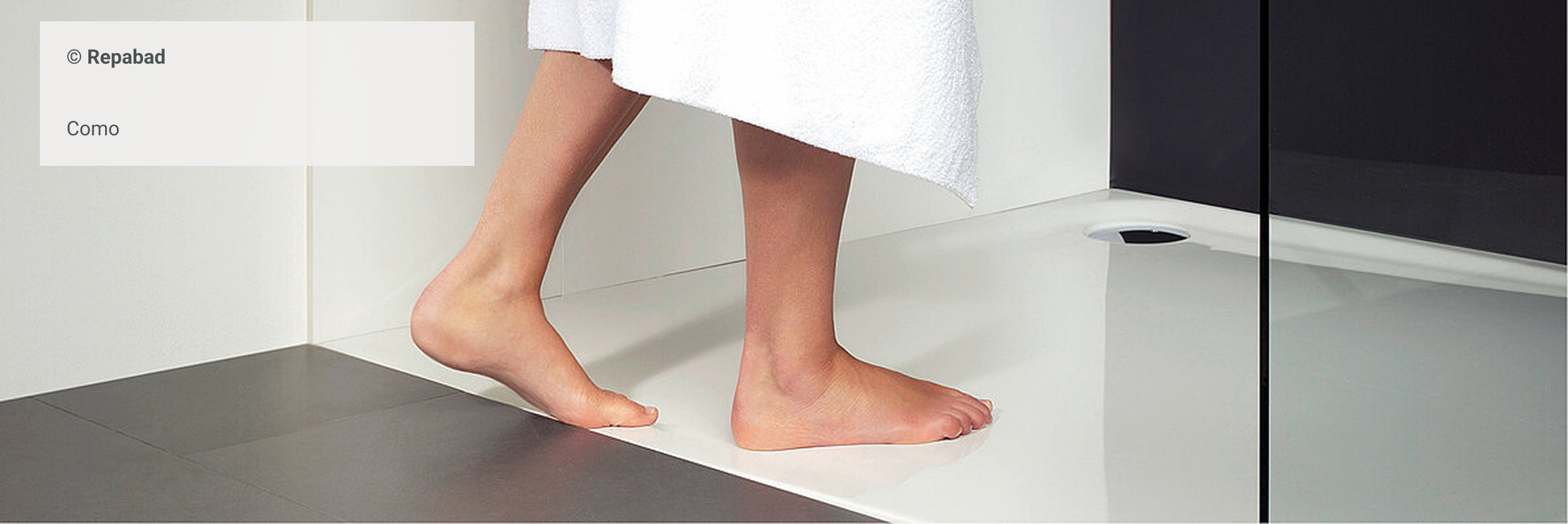 Barfußperson mit weißem Handtuch betritt eine bodenebene Dusche mit glatter, weißer Duschwanne – Modell Como von Repabad