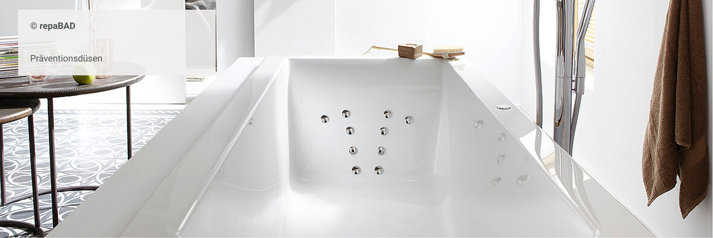 Weiße Badewanne mit integrierten Präventionsdüsen, daneben moderne freistehende Armatur und Handtuch an der Wand