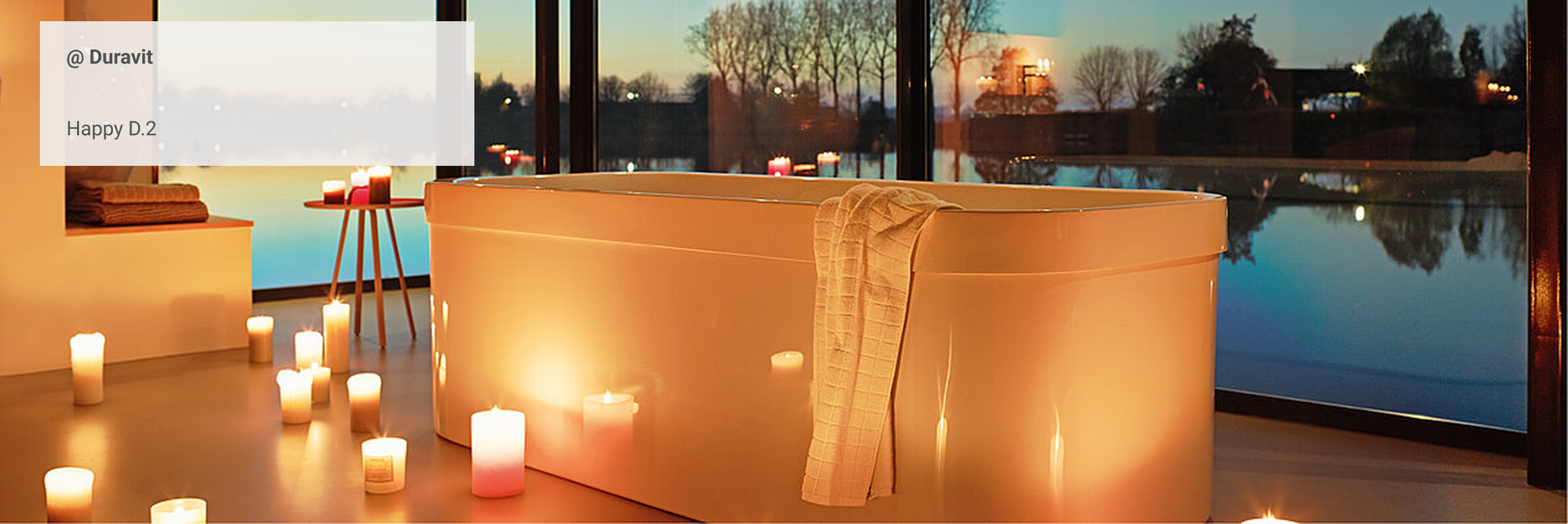 Freistehende Badewanne in stimmungsvoller Abendatmosphäre mit Kerzenlicht, vor großen Fenstern mit Blick auf einen See