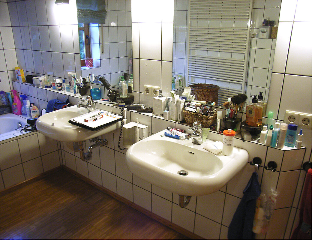 Veraltetes Badezimmer mit beigen Fliesen, kleinem Waschbecken und unordentlicher Ablage vor der Sanierung